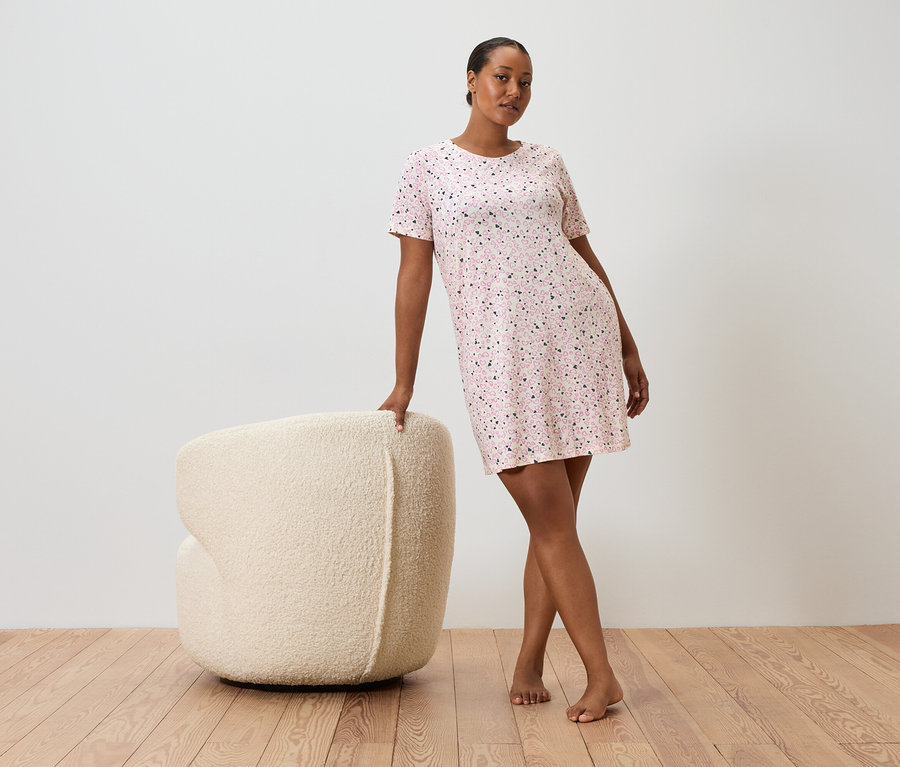 Kobieta opiera się o kremowy fotel, ma na sobie różową sukienkę w kropki z krótkim rękawem.