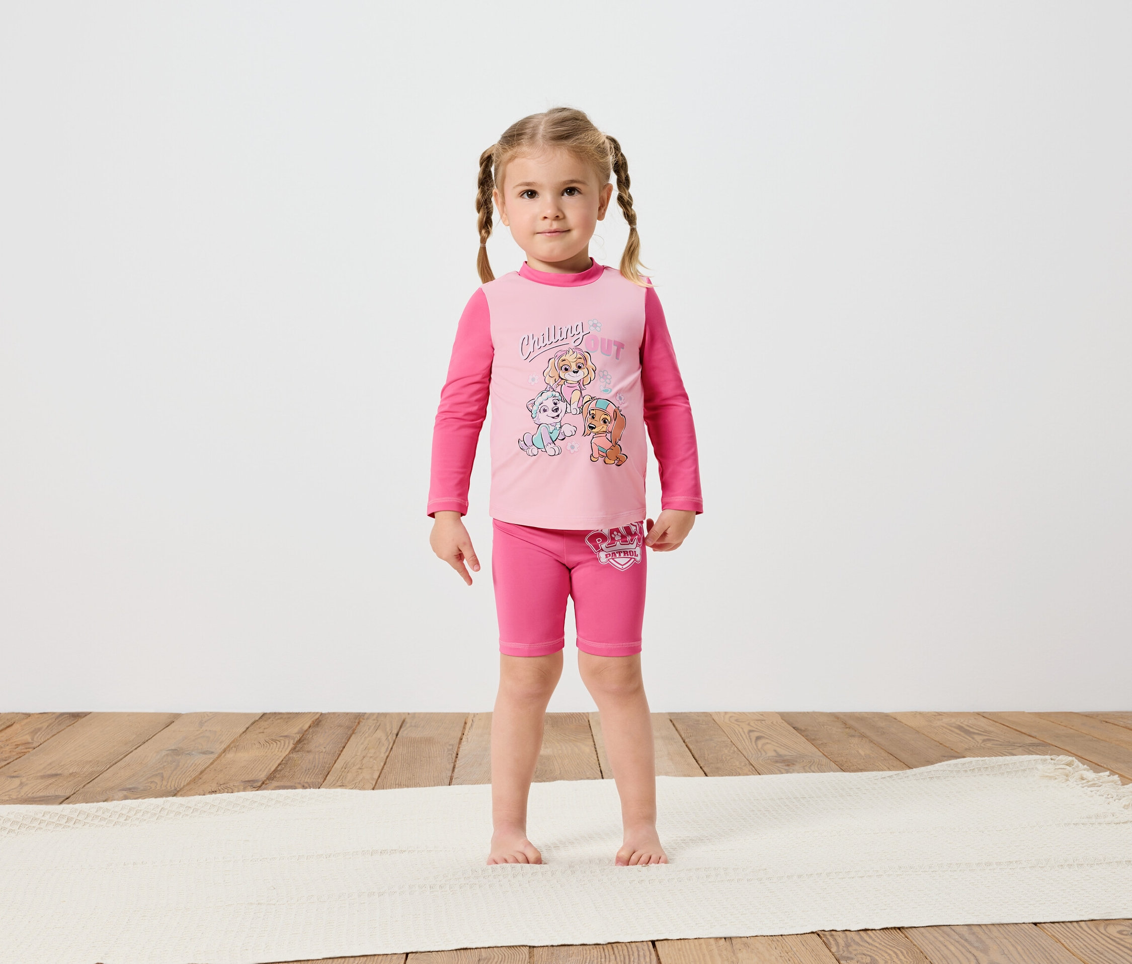 Mała dziewczynka z warkoczami stoi boso na kremowym dywanie, ubrana w różową koszulkę z długim rękawem i spodenki Paw Patrol.