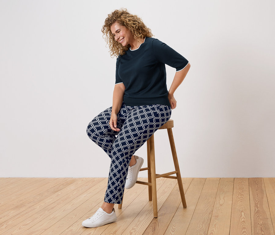 Kobieta o kręconych włosach siedzi na stołku, ubrana w granatową bluzkę i wzorzyste spodnie.