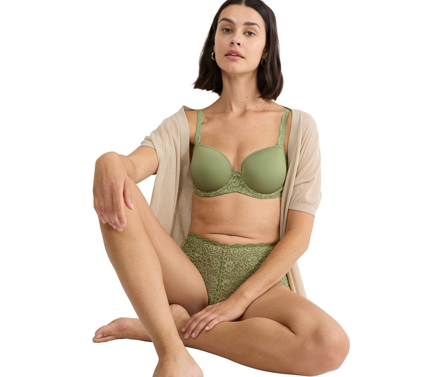 Kobieta siedzi ze skrzyżowanymi nogami, ubrana w zielony biustonosz i zielone koronkowe figi, wraz z beżowym kardiganem.