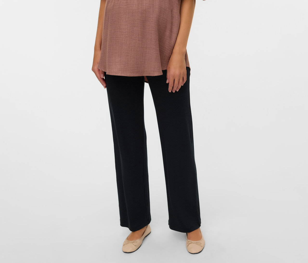 Kobieta ma na sobie brązową bluzkę i czarne legginsy o luźnym kroju MAMALICIOUS „MLEMILY” oraz beżowe baleriny.