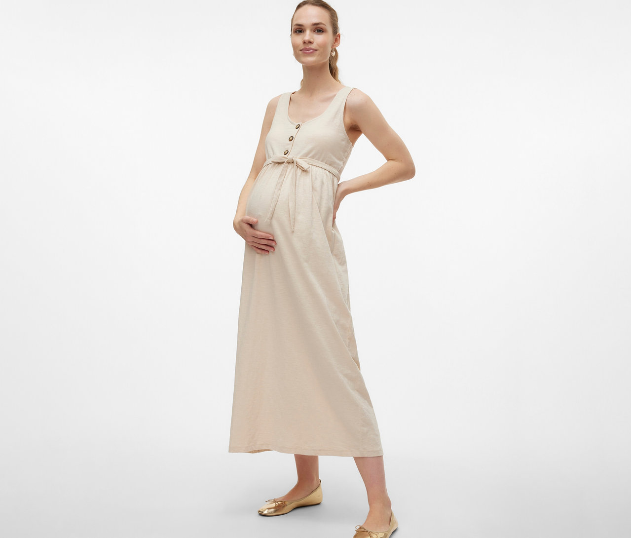 Kobieta w ciąży w sukience ciążowej MAMALICIOUS „MLEVI LIA” z paskiem i guzikami pozuje z rękami na brzuchu i biodrze.