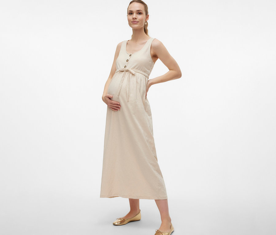 Kobieta w ciąży w sukience ciążowej MAMALICIOUS „MLEVI LIA” z paskiem i guzikami pozuje z rękami na brzuchu i biodrze.