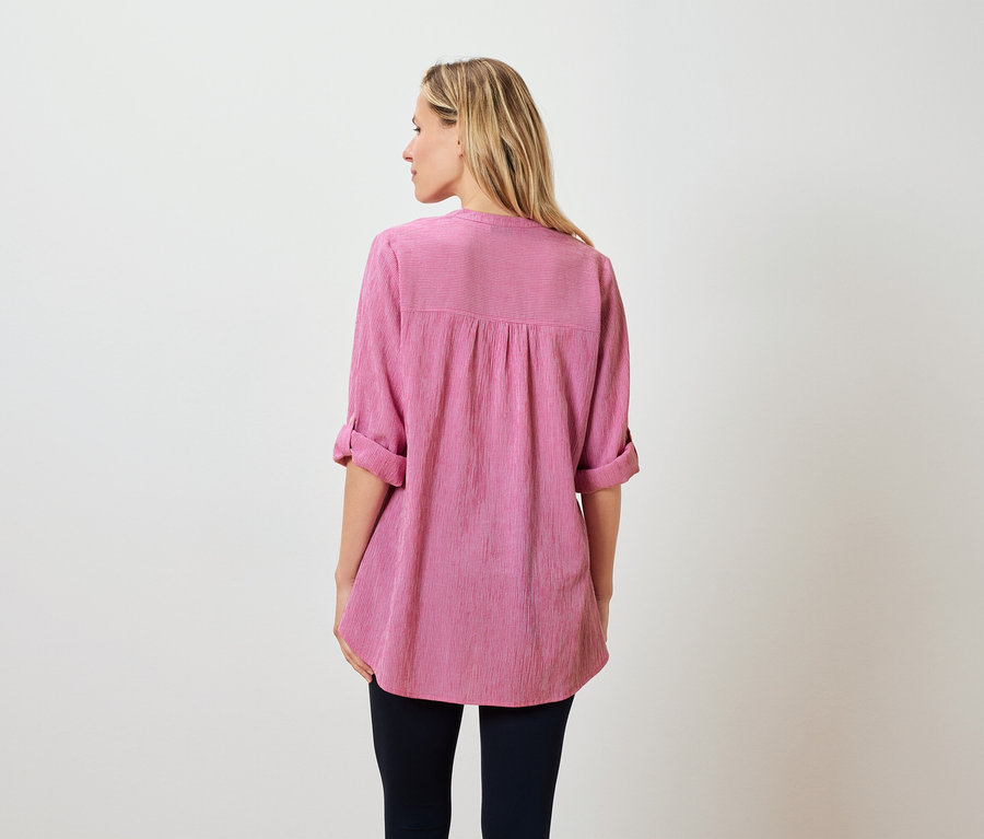 Widok z tyłu kobiety z blond włosami do ramion, ubranej w różową bluzkę i ciemne spodnie.