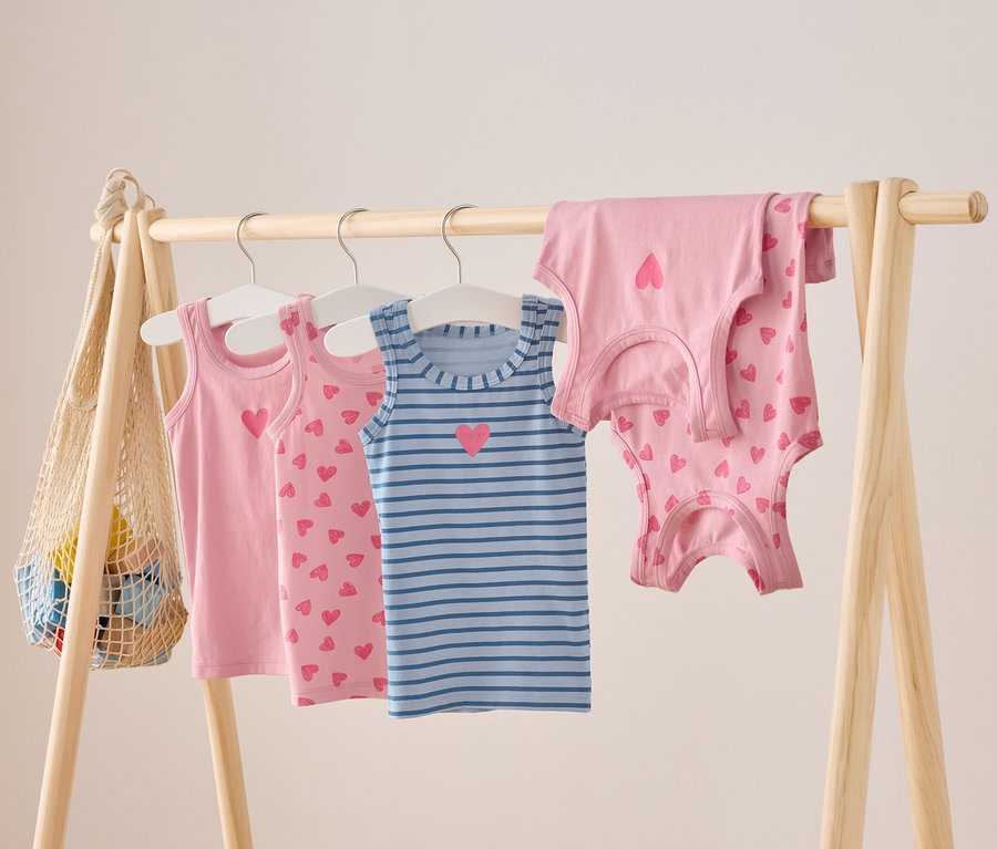Na drewnianym wieszaku wiszą różowe i niebieskie, w paski, dziecięce topy na ramiączkach z motywem serca.