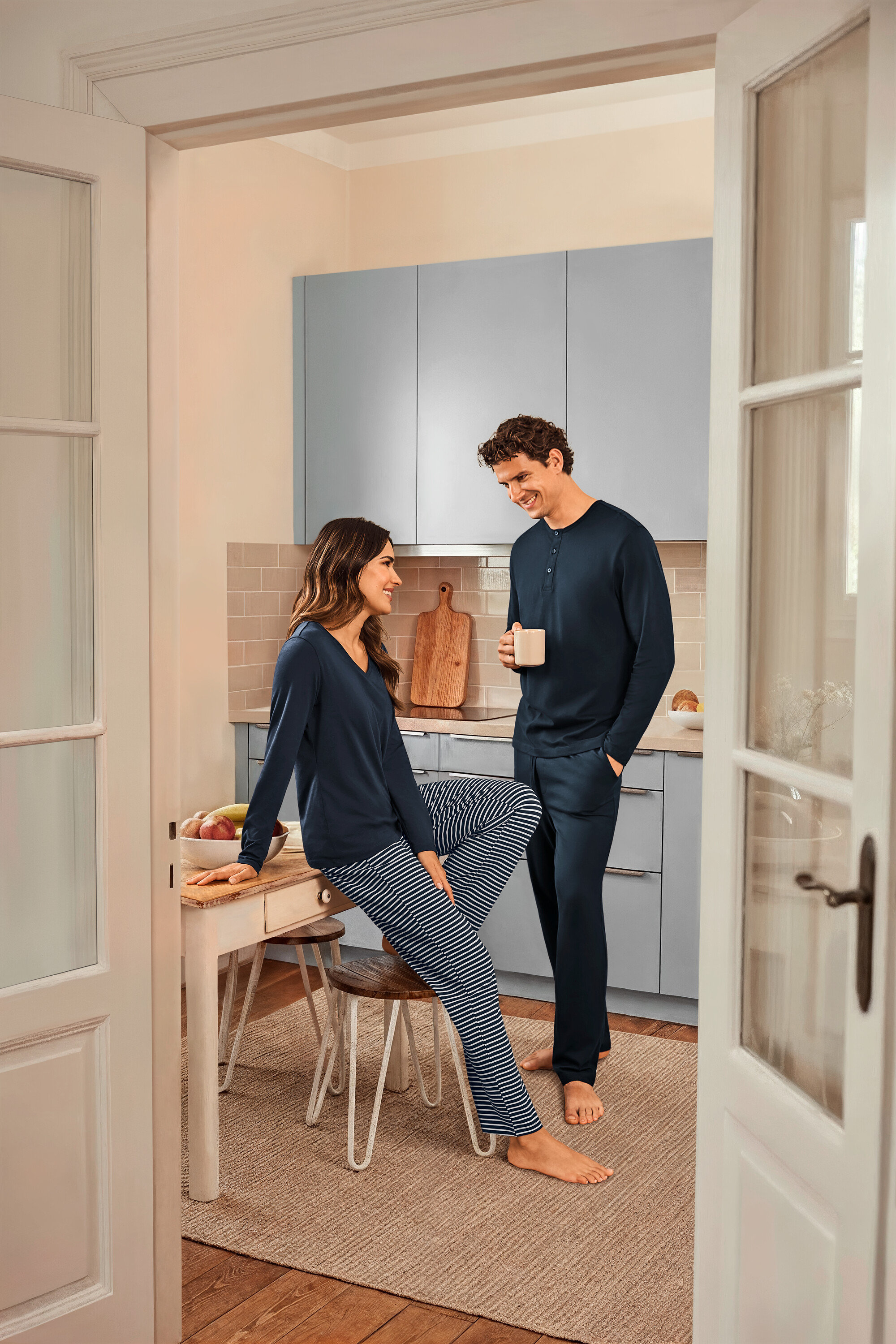 W kuchni stoi kobieta w ciemnoniebiesko-granatowej piżamie w paski i ciemnoniebieskiej górze, siedząca na stole, oraz mężczyzna trzymający kubek i ubrany w ciemnoniebieską piżamę.