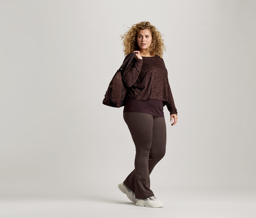 Kobieta z kręconymi włosami ma na sobie brązową bluzkę w zebrę, brązowe legginsy i buty sportowe. Ma brązową torbę na ramieniu.