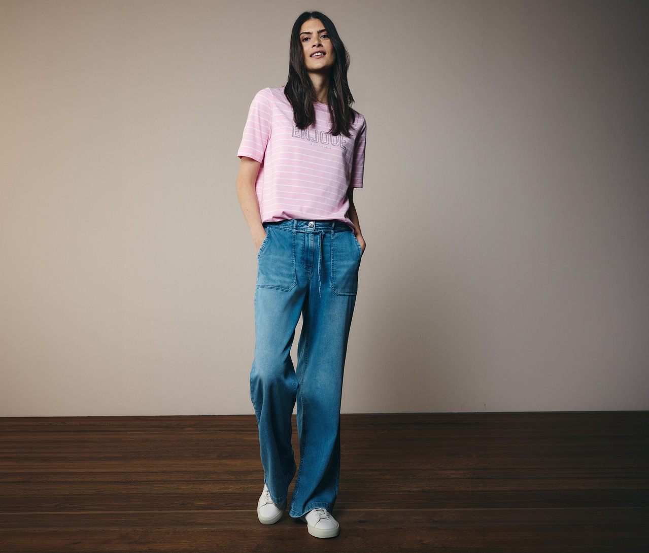 Kobieta w koszulce CECIL w różowe paski i szerokich dżinsach oraz białych tenisówkach pozuje z rękami w kieszeniach.