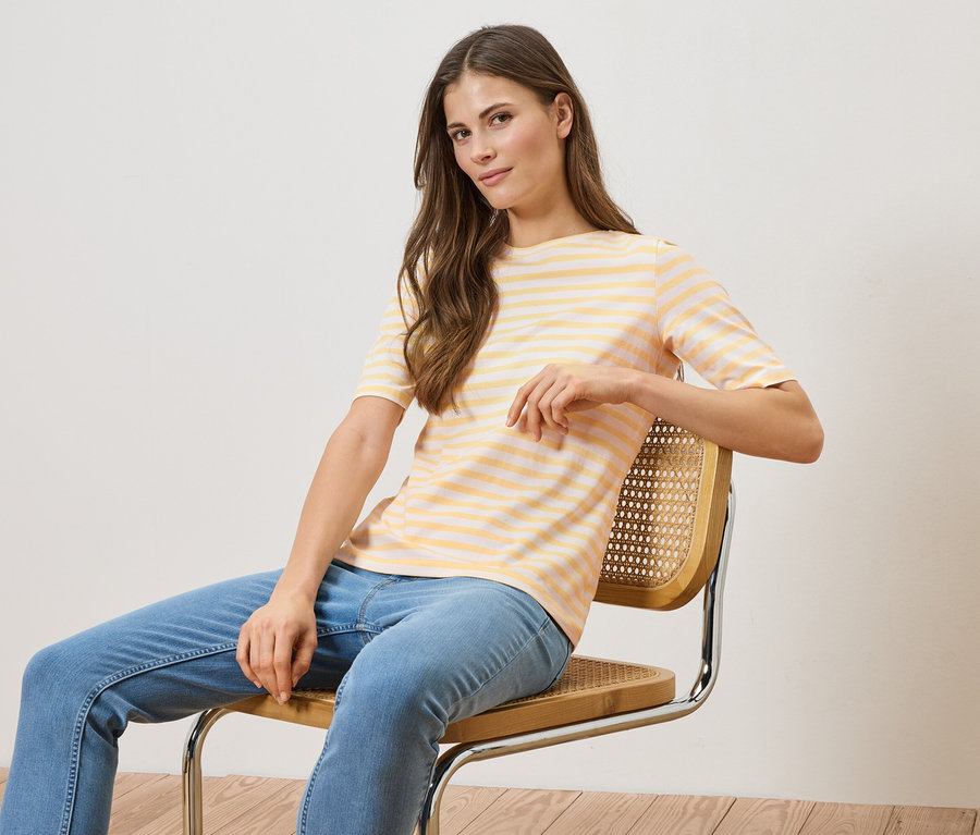 Kobieta siedzi na krześle, ma na sobie bluzkę w żółto-białe paski i niebieskie dżinsy.