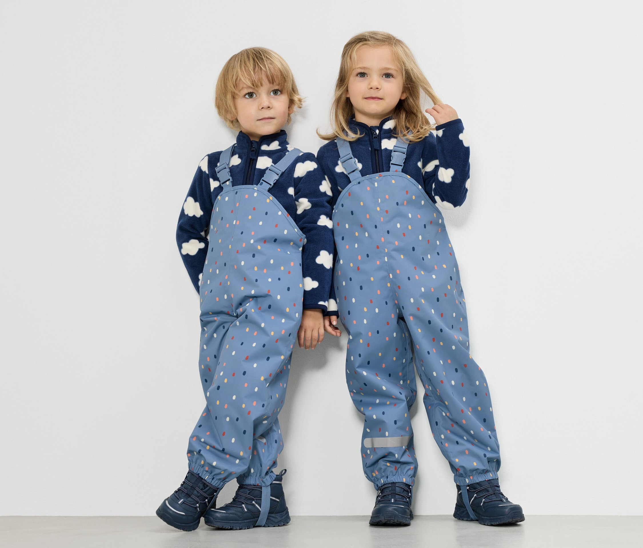 Dwoje dzieci stoi obok siebie, chłopiec i dziewczynka, oboje ubrani w niebieskie bluzy polarowe z motywem chmur i niebieskie ogrodniczki przeciwdeszczowe w kolorowe kropki.