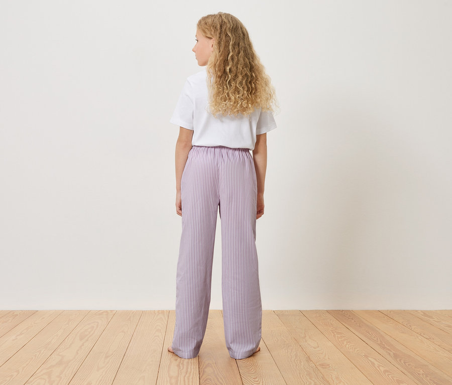 Widok z tyłu młodej dziewczyny z blond, kręconymi włosami, ubranej w białą koszulkę i jasnofioletowe spodnie w paski.