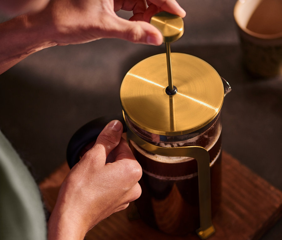 Osoba naciska na złoty zaparzacz tłokowy o pojemności 800 ml na 6 filiżanek, umieszczony na drewnianej podstawie.