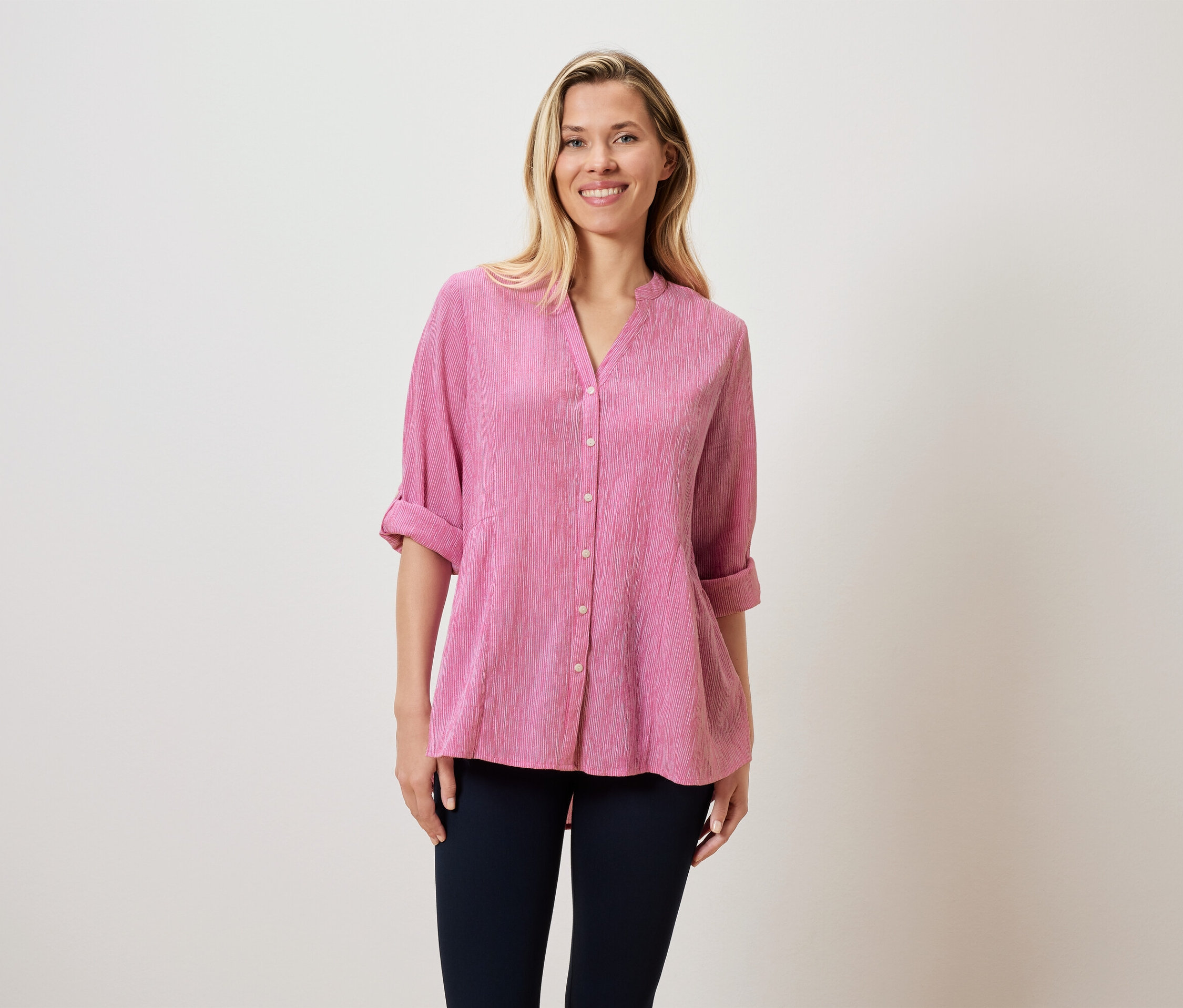 Kobieta o blond włosach ma na sobie różową bluzkę i ciemne spodnie.