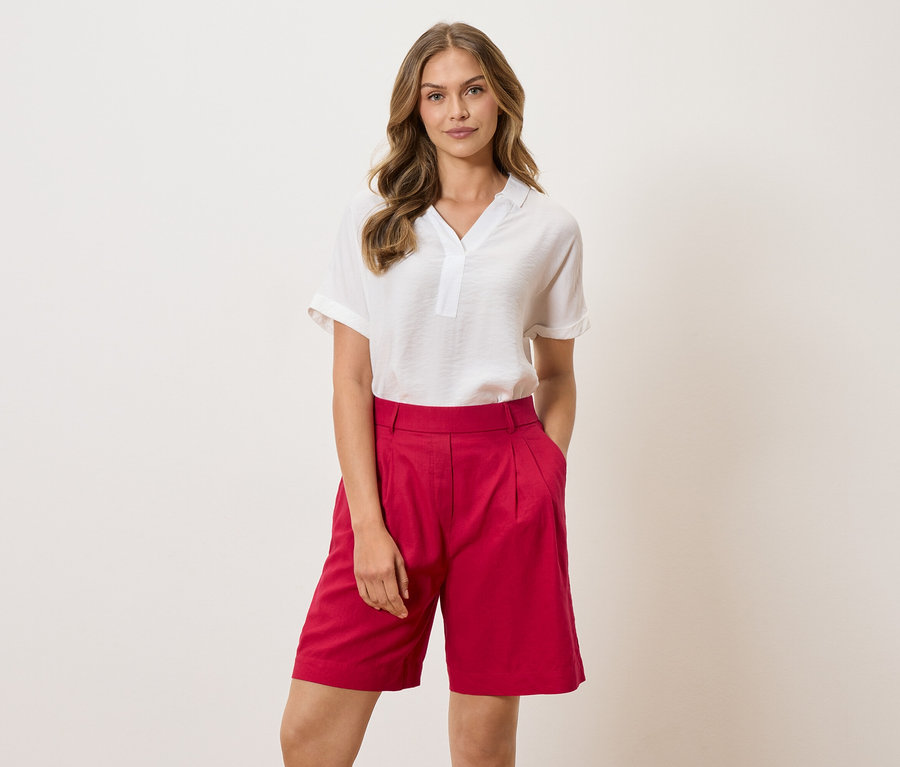 Kobieta w białej bluzce i czerwonych szortach stoi na jasnym tle.