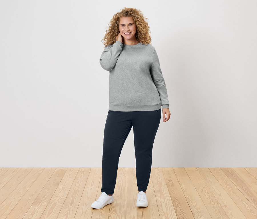 Kobieta z kręconymi włosami w szarym swetrze i granatowych spodniach.
