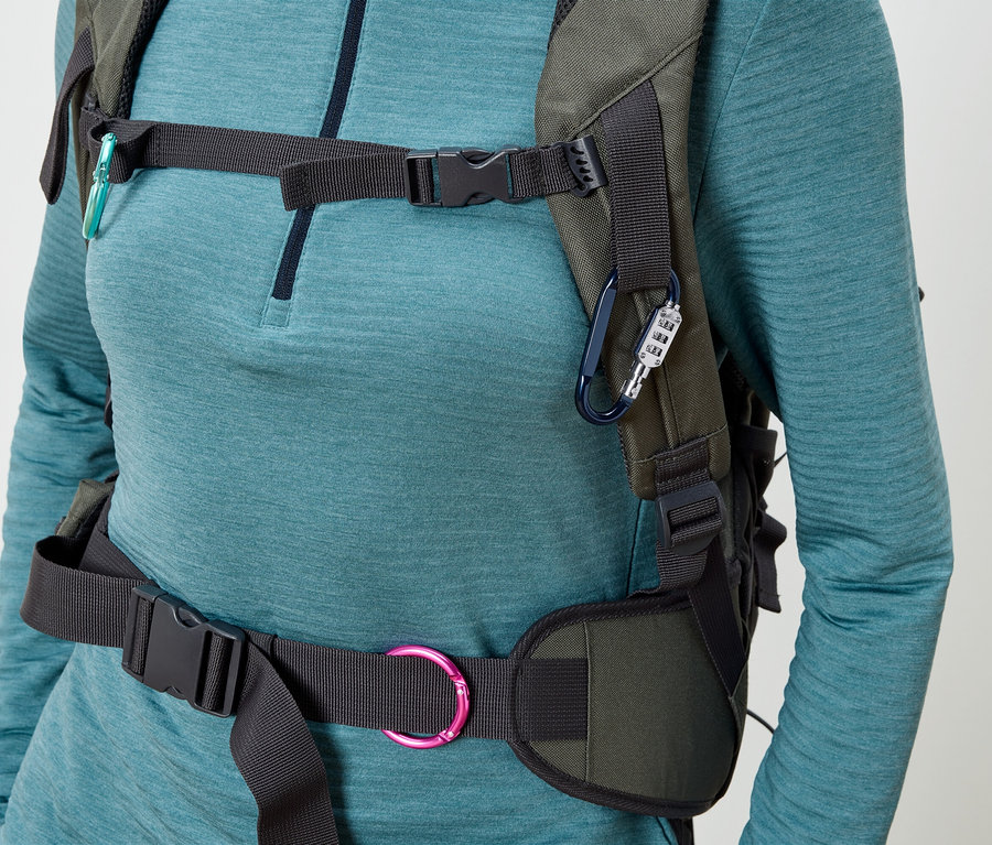 Kobieta ma na sobie oliwkowozielony plecak z czarnymi paskami i klamrami, przymocowany niebieskim karabińczykiem z zamkiem szyfrowym i turkusowym karabińczykiem.