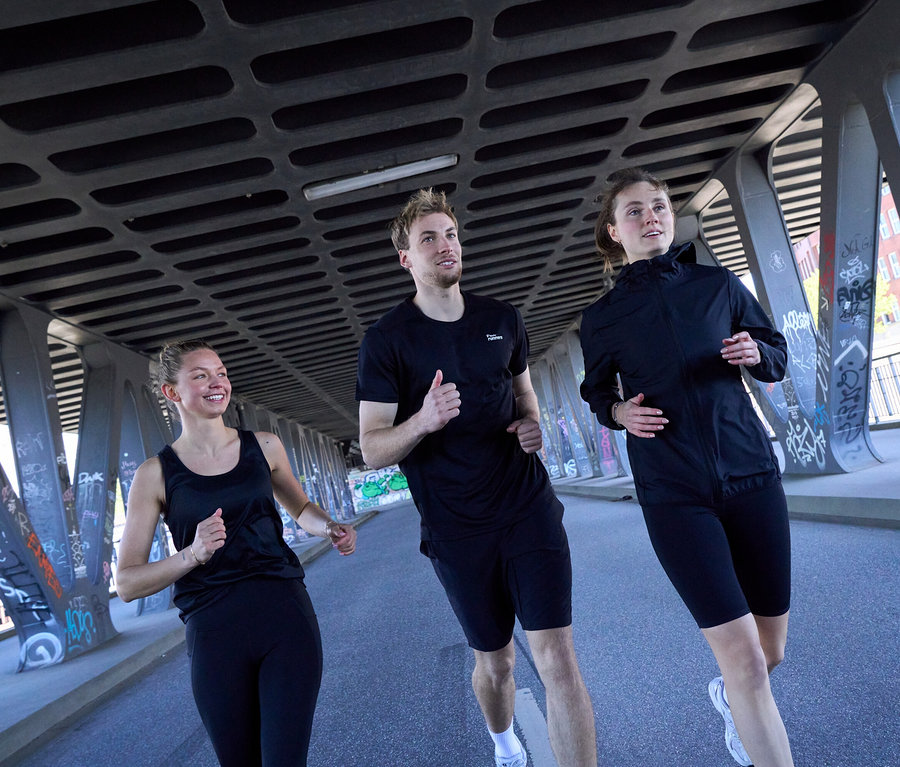 Trzy osoby biegną pod mostem: Kobieta w czarnym topie sportowym i legginsach sportowych „ActivePower”, mężczyzna w czarnej koszulce sportowej i czarnych szortach sportowych, oraz kobieta w czarnej kurtce wiatrówce „packable” i czarnych kolarkach „ActivePower”.