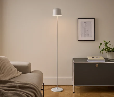 Nowoczesny salon z beżową sofą, białą lampą podłogową i ciemnoszarą szafką. Oświetlona lampa stoi między sofą a szafką, na której leżą książki i doniczkowa roślina pod oprawionym obrazem.