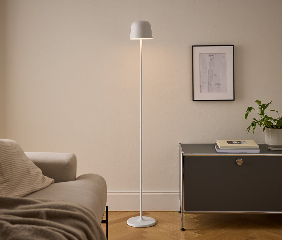 Nowoczesny salon z beżową sofą, białą lampą podłogową i ciemnoszarą szafką. Oświetlona lampa stoi między sofą a szafką, na której leżą książki i doniczkowa roślina pod oprawionym obrazem.