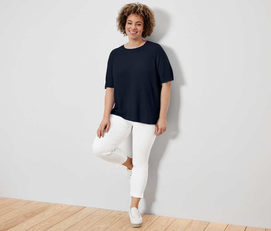 Kobieta oparta o ścianę ma na sobie granatową bluzkę z cienkiej dzianiny, białe spodnie i tenisówki.