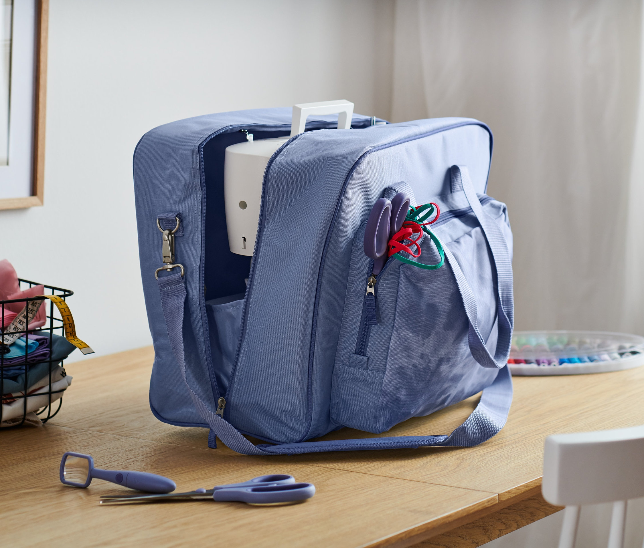 Niebieska torba na maszynę do szycia otwarta na drewnianym stole, w tle kosz z materiałami i okno.