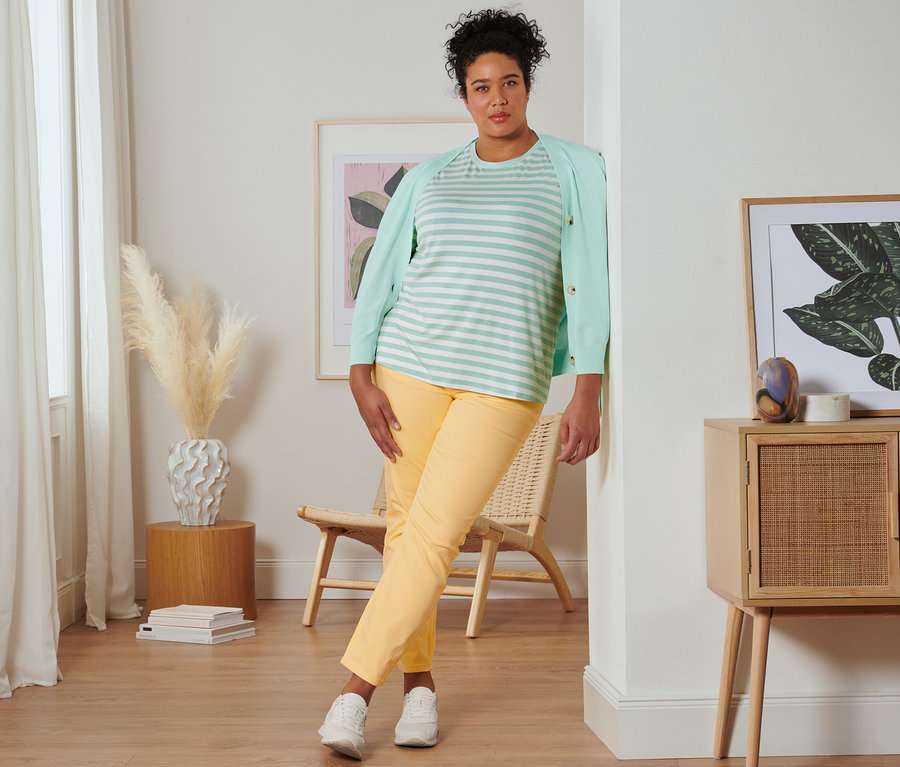 Kobieta oparta o ścianę ma na sobie miętową koszulkę w paski, kardigan z dekoltem w serek, żółte spodnie dżinsowe o kroju slim fit i buty typu sneakers ze skóry welurowej.