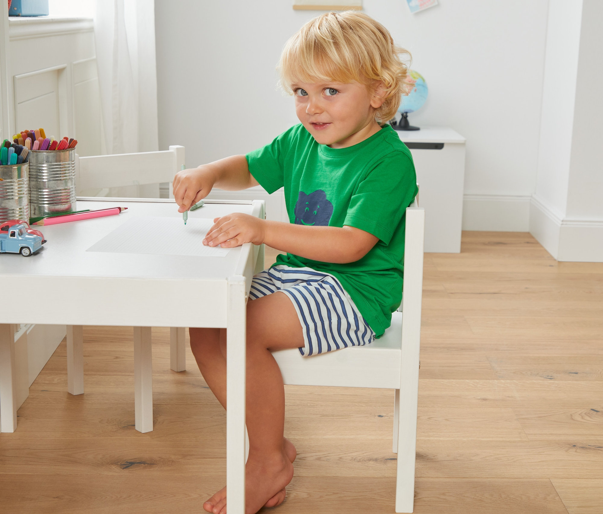 Chłopiec siedzi przy stole i rysuje na kartce. Ma na sobie zieloną koszulkę i Piżamy z koszulką i spodenkami z bawełny ekologicznej, 2 sztuki.