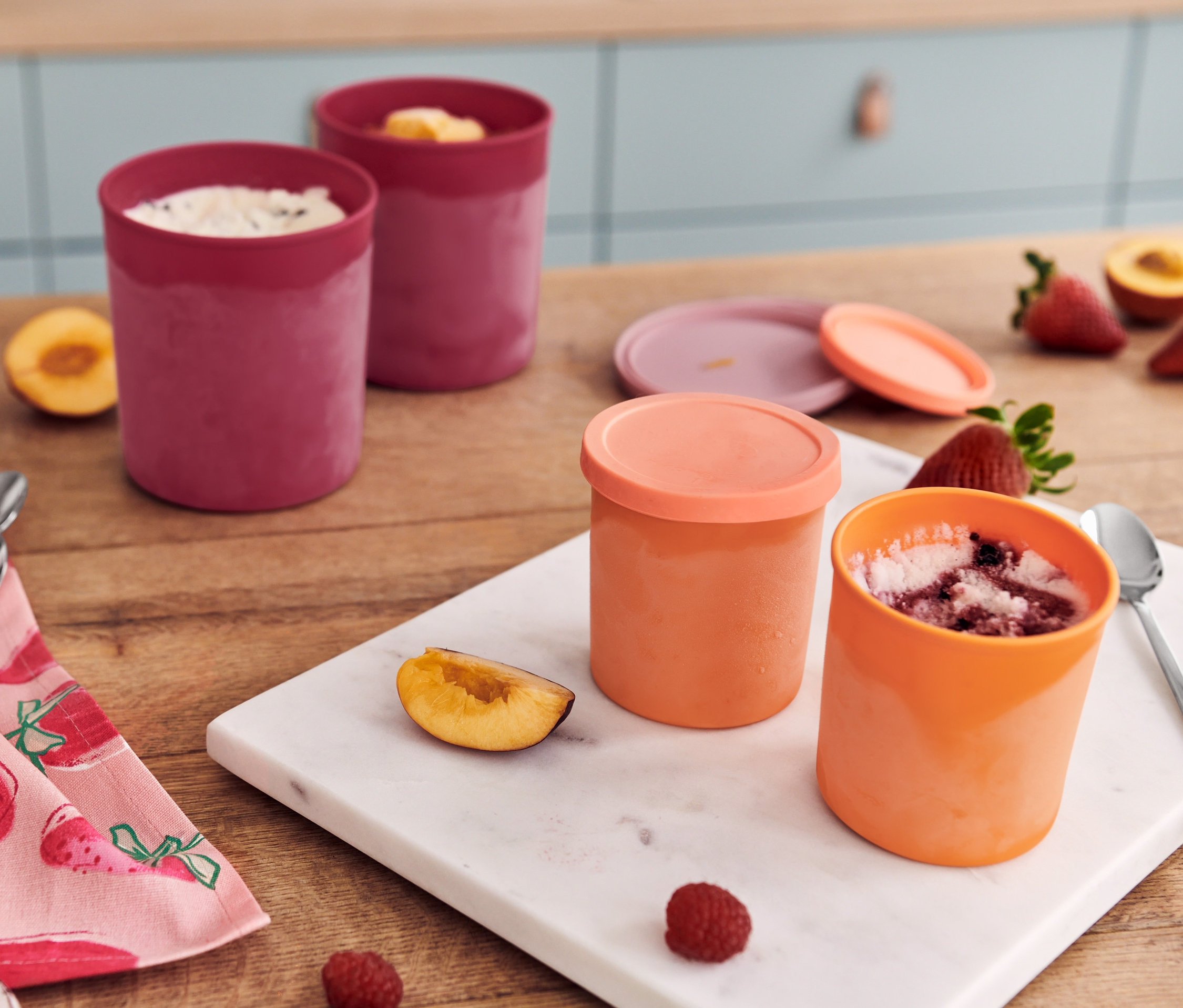 Dwa czerwone i dwa pomarańczowe pojemniki do mrożenia z owocami i łyżeczkami na stole ze ścierką.