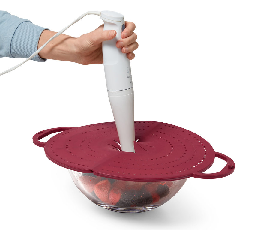 Ręka trzyma blender zanurzeniowy nad miską z owocami, na której znajduje się czerwona osłona na patelnię 4 w 1.