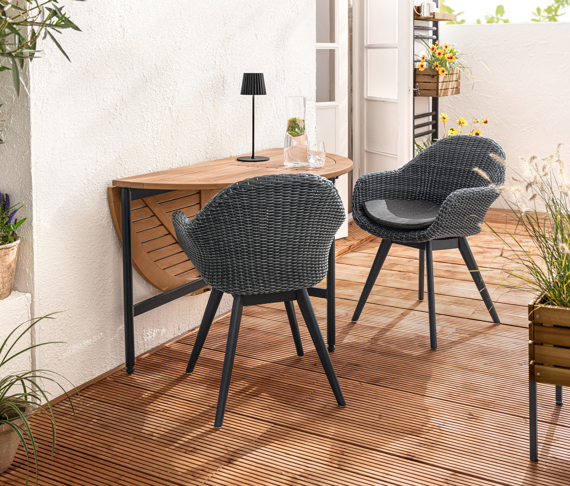 Dwa krzesła muszelki z plecionką polirattanową stoją przy rozkładanym stole „Elin”. Na stole stoi akumulatorowa lampa stołowa LED do użytku na zewnątrz, czarna. Na pierwszym planie skrzynka na rośliny „Elin”.