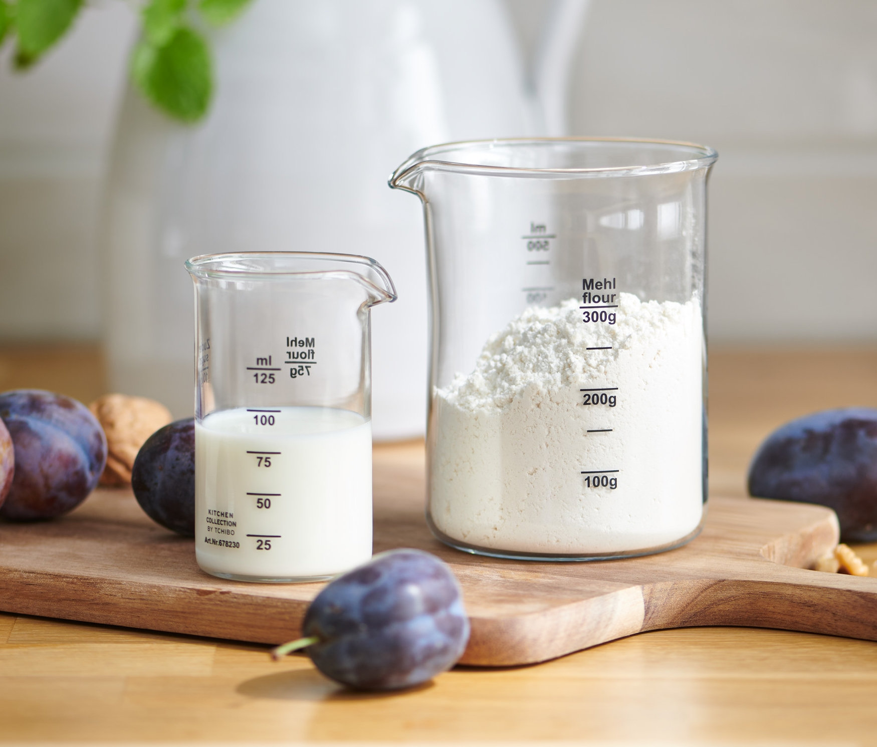 Dwa kubki-miarki, jeden z mlekiem, drugi z mąką, leżą na drewnianej desce razem ze śliwkami i orzechami.