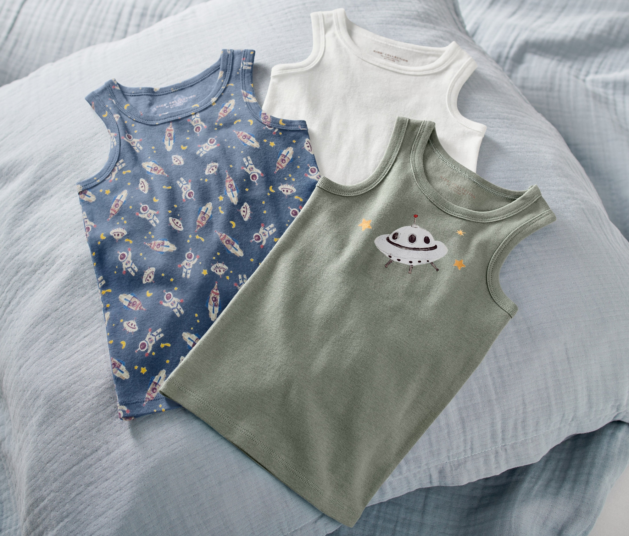 Na jasnoniebieskiej poduszce leżą trzy podkoszulki dziecięce: niebieski we wzory z motywem kosmosu, zielony z nadrukiem UFO i biały.