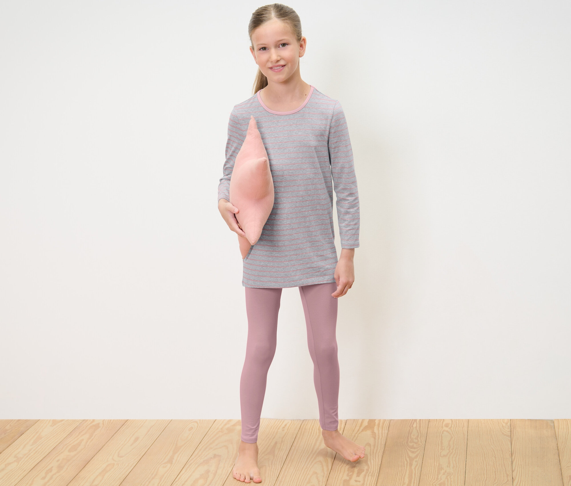 Dziewczyna trzyma różową poduszkę i ma na sobie Piżamy, 2 zestawy.