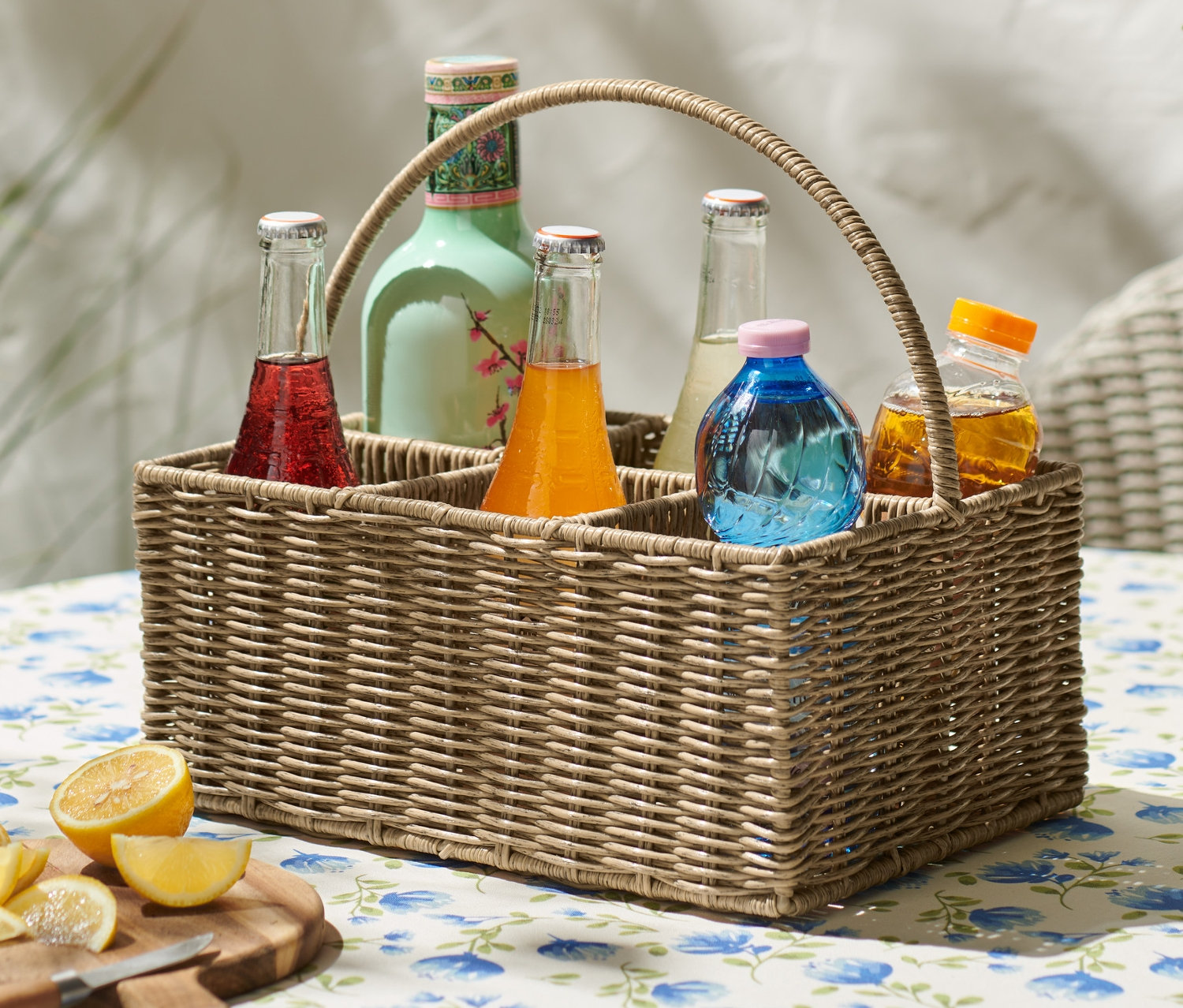 Wiklinowy kosz na butelki z kolorowymi napojami na stole z kwiatowym obrusem.