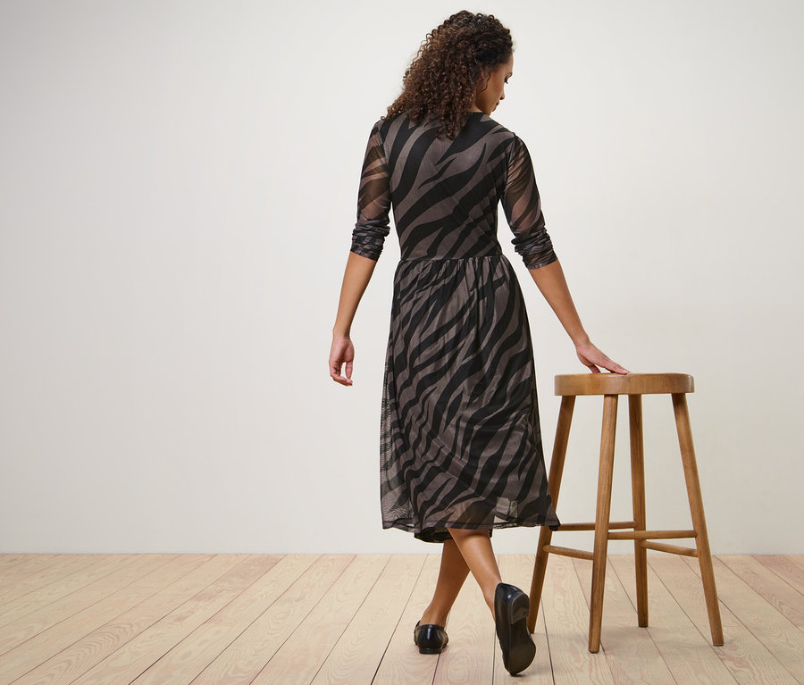 Kobieta pozuje w sukience midi i opiera rękę o drewniany stołek.