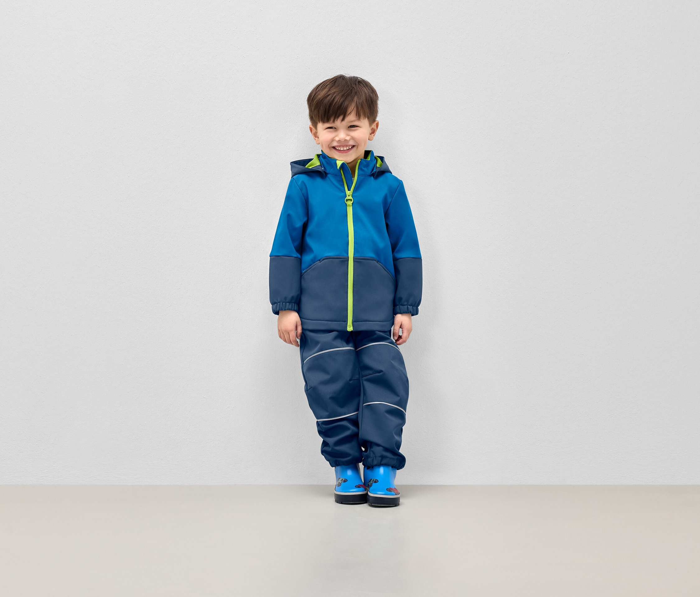 Uśmiechnięty chłopiec pozuje w niebieskiej kurtce z kapturem i granatowych spodniach z odblaskowymi paskami.
