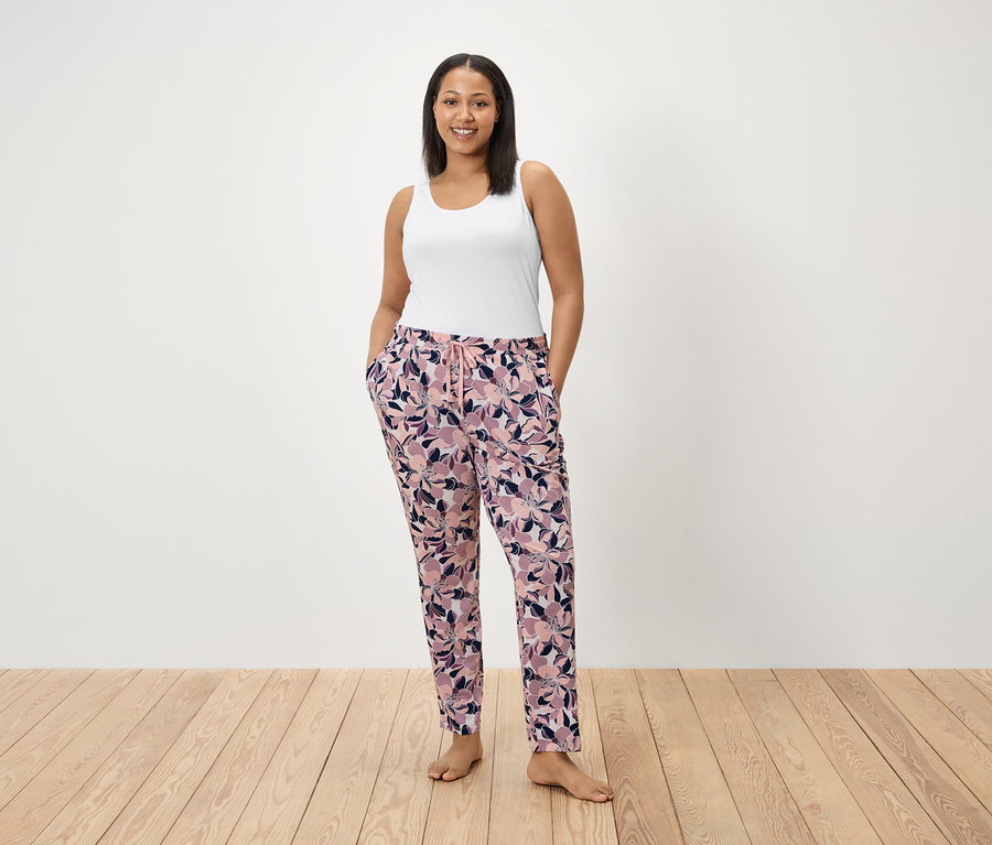 Kobieta stoi z rękami w kieszeniach spodni w kwiatowy wzór. Ma na sobie biały podkoszulek.