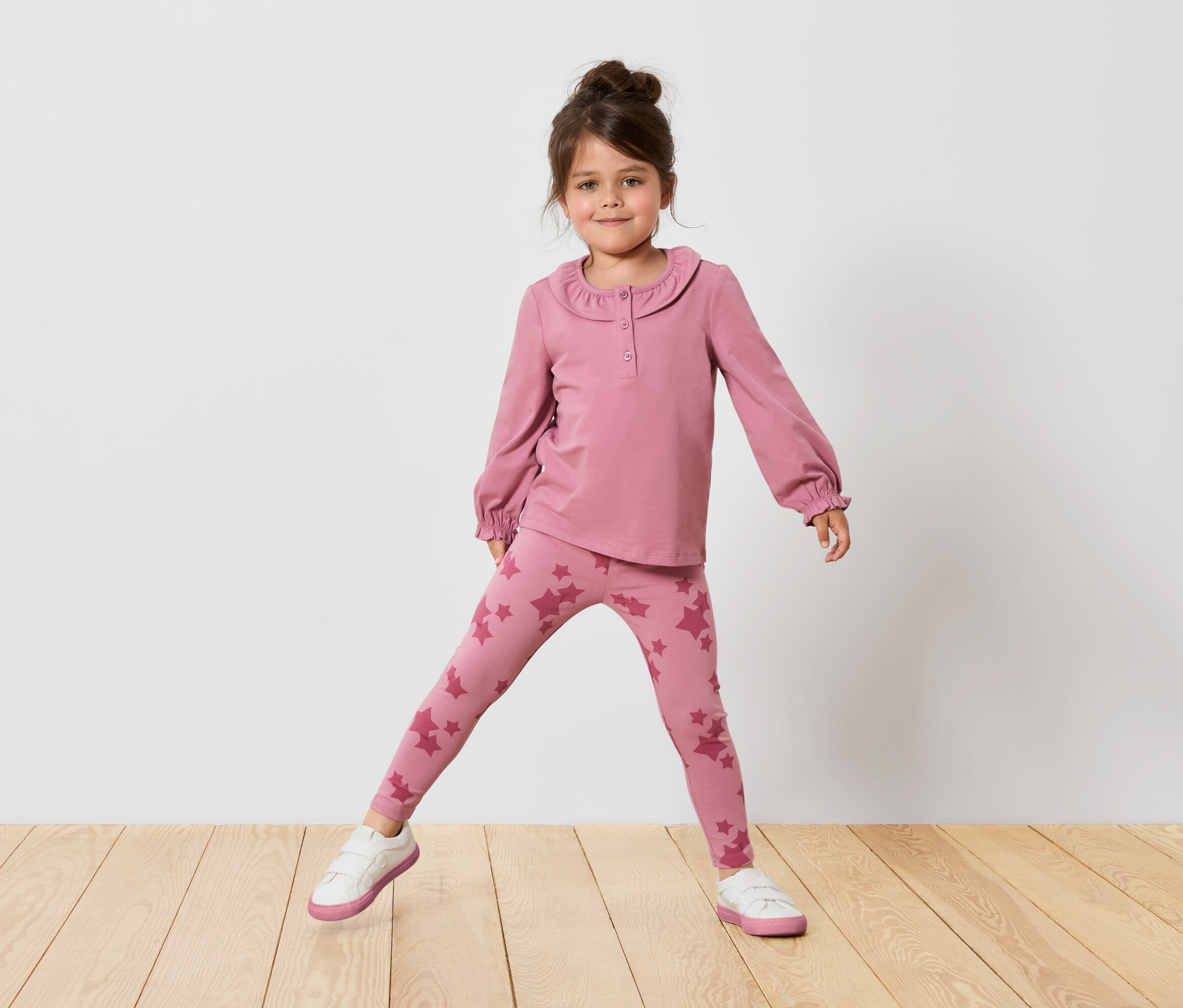Dziewczynka w różowej bluzce z długim rękawem i legginsach w gwiazdy stoi na drewnianej podłodze przed białą ścianą.
