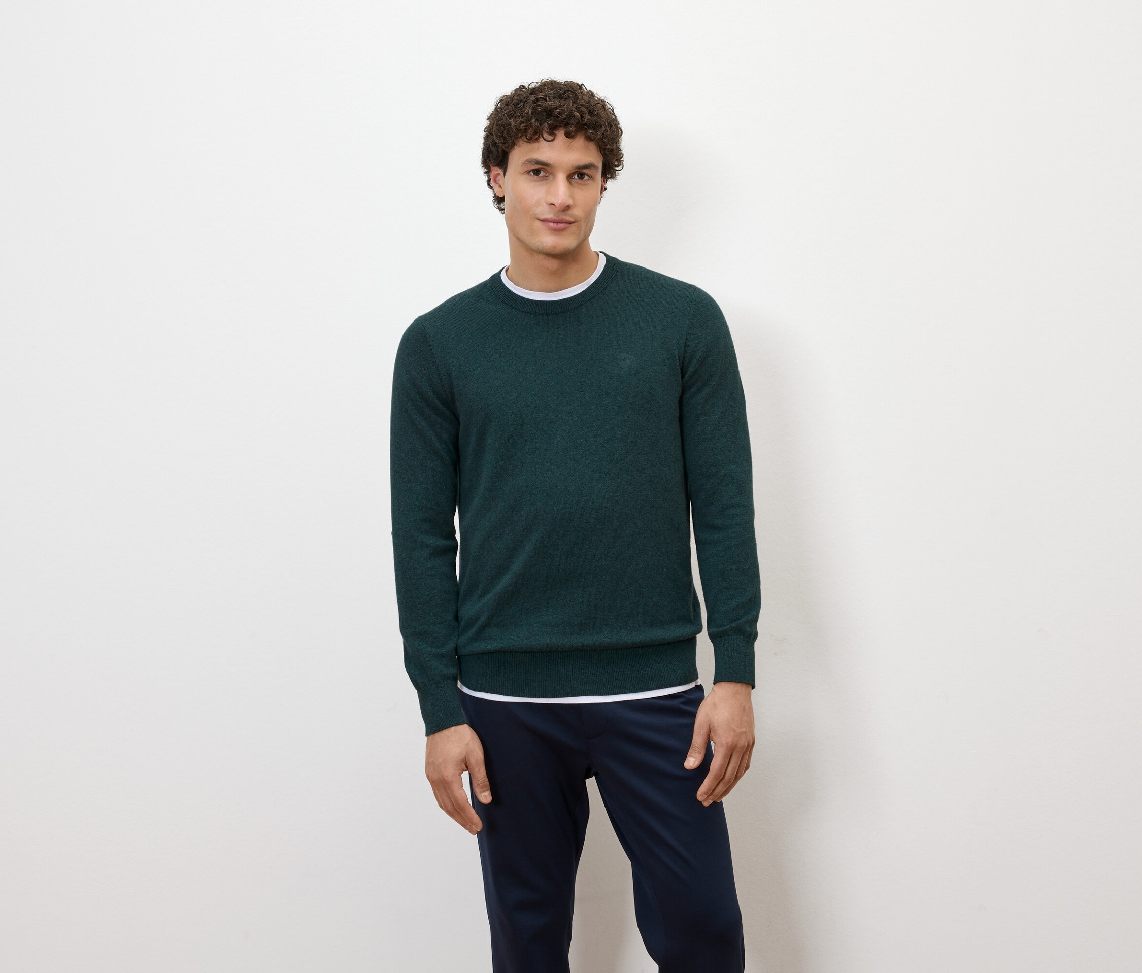 Mężczyzna pozuje na białym tle w ciemnozielonym swetrze i granatowych spodniach.