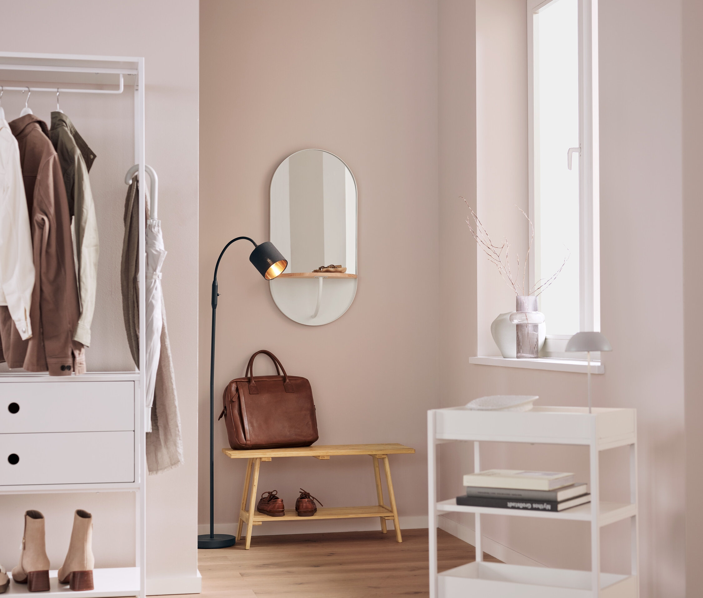 We wnętrzu znajduje się biały regał z ubraniami, owalne lustro, brązowa skórzana torba na ławce, czarna lampa i biały wózek na kółkach.