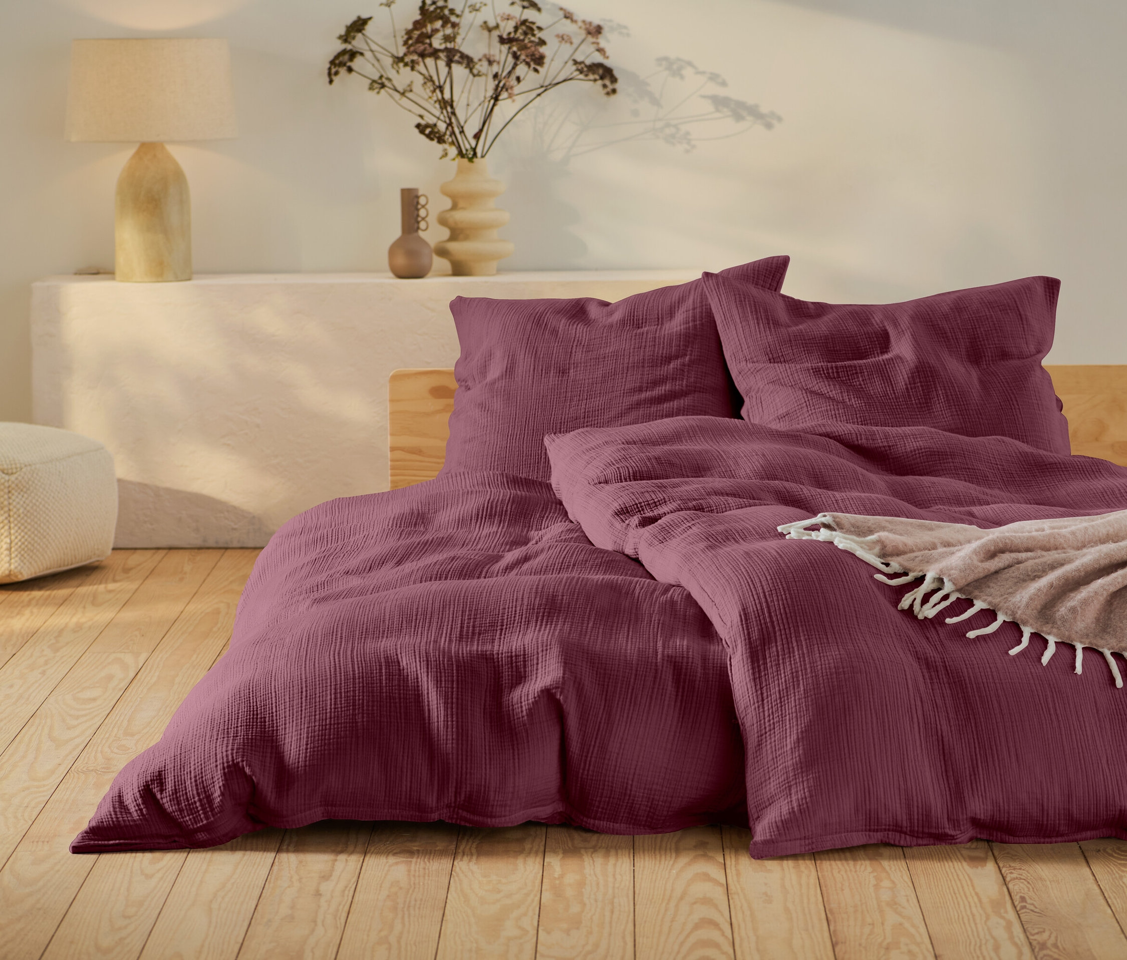 Burgundowa pościel na łóżku z drewnianą ramą, beżowy puf i lampa na białym stoliku.