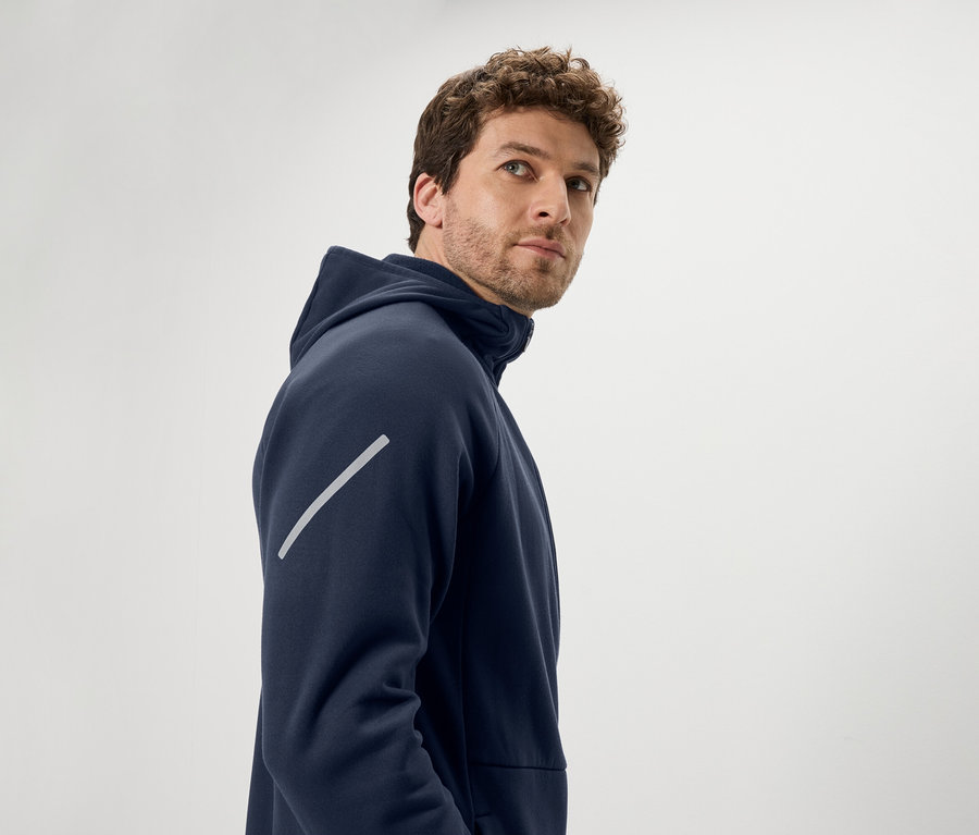 Mężczyzna z brązowymi kręconymi włosami spogląda przez ramię. Ma na sobie granatową bluzę z kapturem i ukośny odblaskowy pasek na ramieniu.