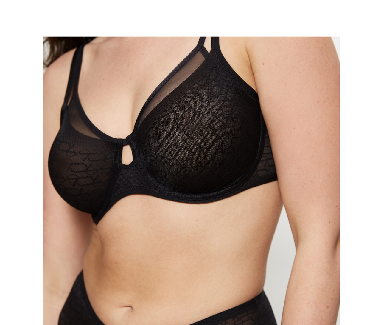 Kobieta w czarnym biustonoszu Triumph Signature Sheer Minimizer i pasujących, wzorzystych majtkach. Biustonosz ma małe wycięcie między miseczkami.