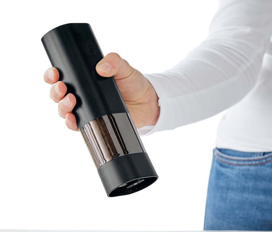 Ręka trzyma czarny młynek do przypraw nad białym stołem, z którego wysypują się przyprawy.