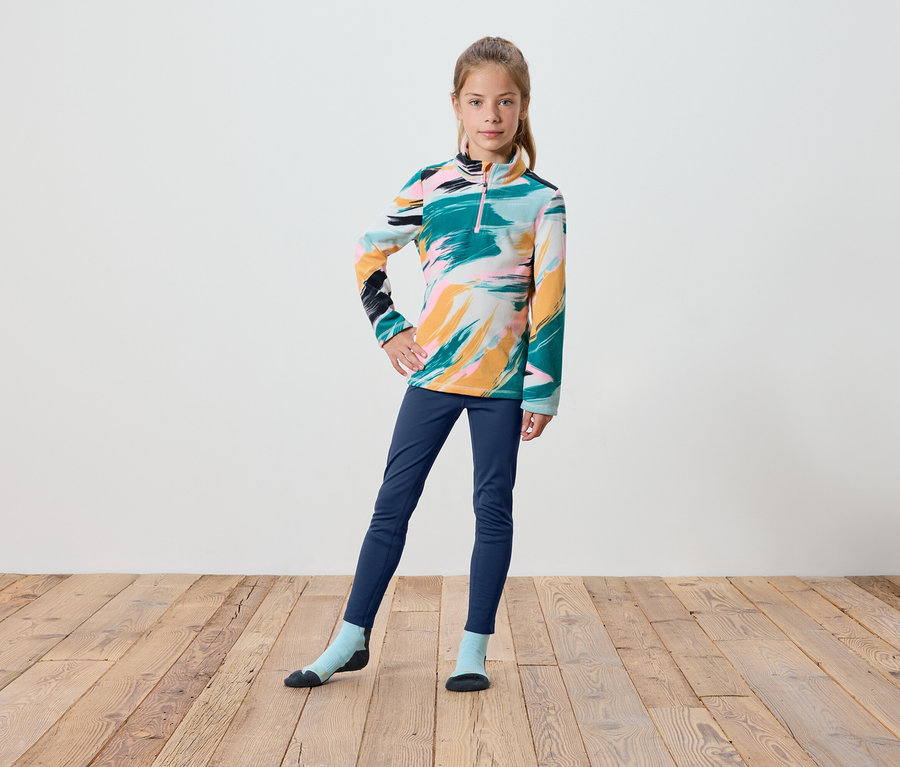 Dziewczyna w kolorowej bluzie z długim rękawem, granatowych spodniach i skarpetkach stoi na drewnianej podłodze.