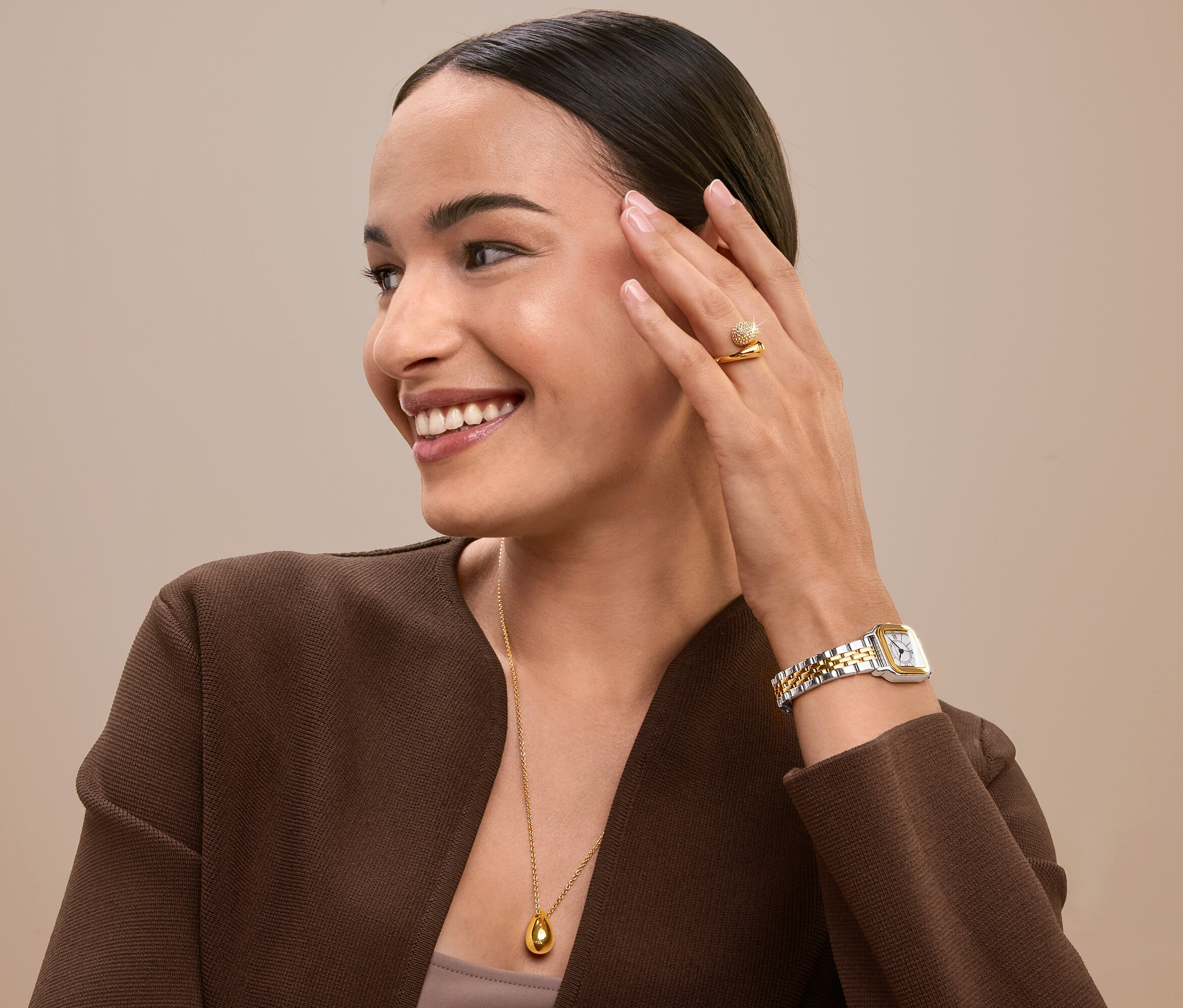Uśmiechnięta kobieta dotyka twarzy, ma na sobie złoty pierścionek, zegarek i naszyjnik.
