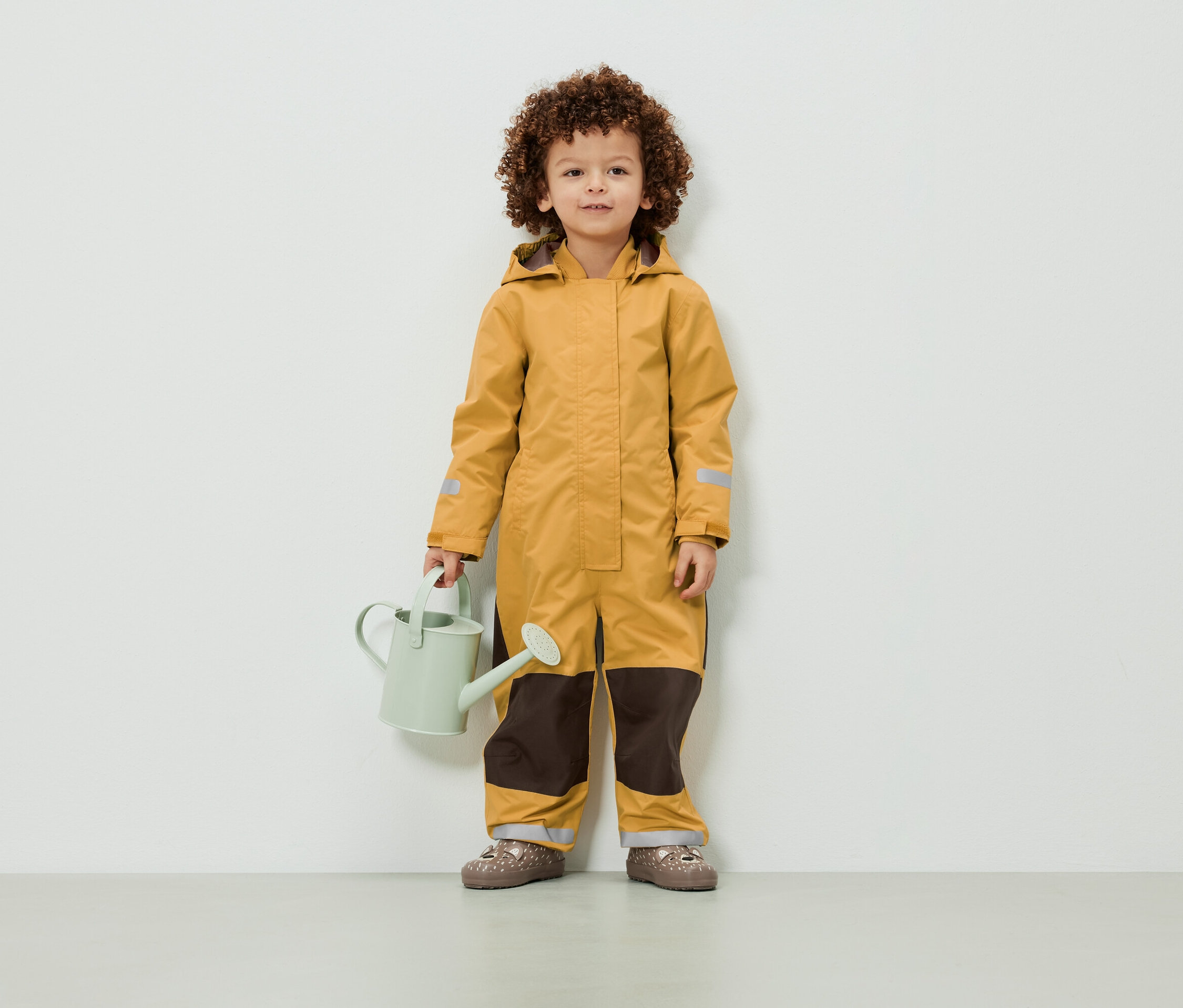 Dziecko z kręconymi włosami stoi w żółtym kombinezonie i trzyma jasnozieloną konewkę.