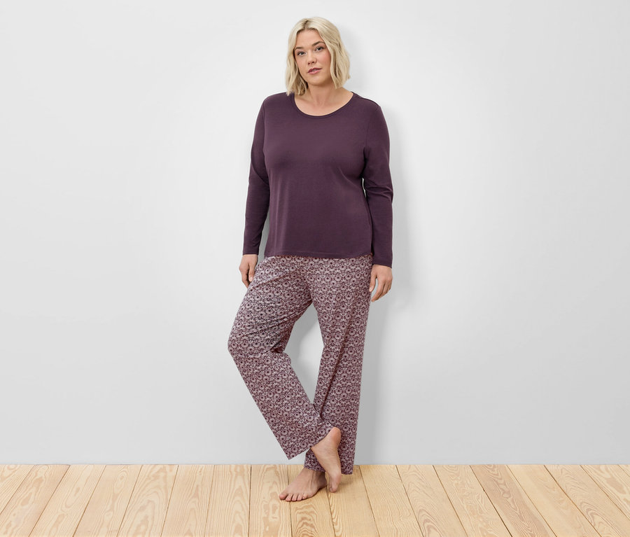 Blondynka w bordowej piżamie z długim rękawem i kwiecistych spodniach pozuje na drewnianej podłodze przy ścianie.