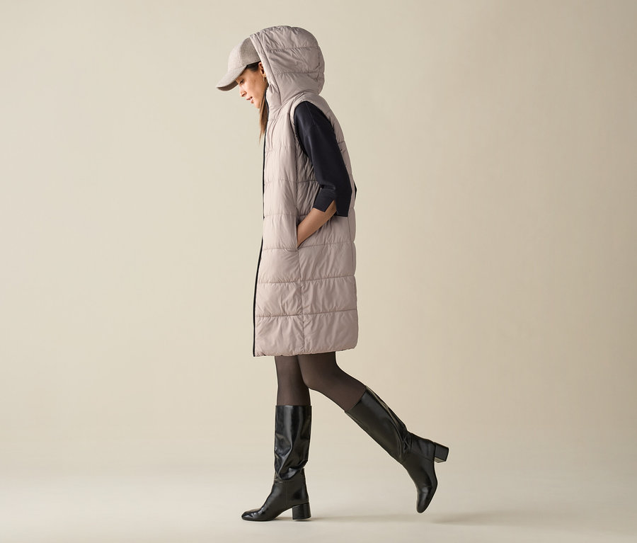 Kobieta idzie, ubrana w pikowaną kamizelkę z kapturem, czapkę i kozaki.