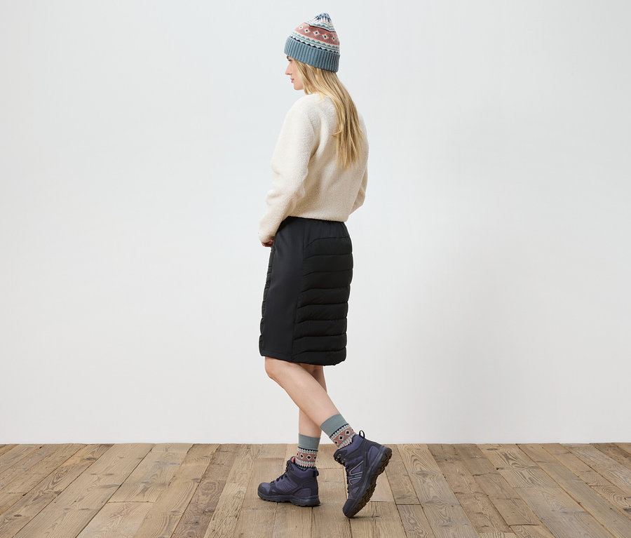 Kobieta pozuje w kremowym swetrze, czarnej spódnicy, butach i dzianinowej czapce.